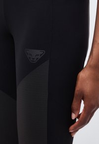 Κοντινό πλάνο ενός χεριού που ακουμπά δίπλα σε μαύρο αθλητικό κολάν με ένα διακριτικό σκοτεινό λογότυπο πάνθηρα στον μηρό.