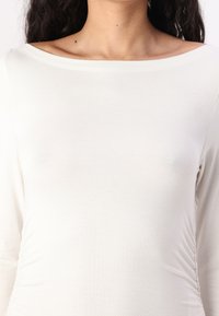 Long-sleeve, cream-colored top made of soft, stretchy fabric with a wide neckline and gathered details on the sides.