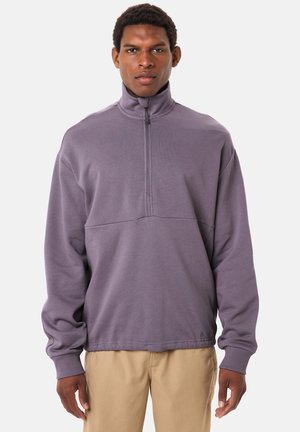 Man die een paarse trui met lange mouwen en een kwart-ritssluiting draagt, samen met een beige broek, staand tegen een effen lichte achtergrond.