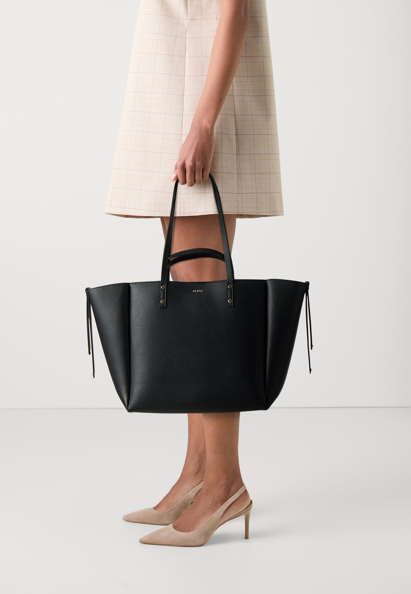 Černá kožená taška přes rameno se strukturovaným designem, dlouhými uchy a decentními zlatými akcenty, umístěná na světlém pozadí.