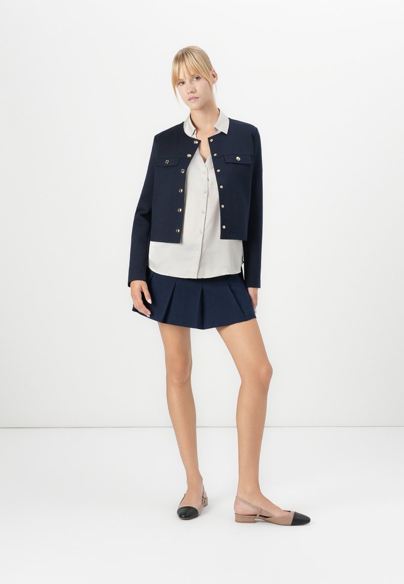 Femme portant une veste courte navy, une chemise beige à boutons, une jupe mini plissée navy et des ballerines beiges avec des bouts noirs sur un fond blanc.