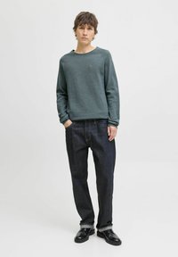 Pulover verde închis cu guler rotund și mâneci raglan, asortat cu blugi închis la culoare. Modelul stă cu mâinile în buzunare.