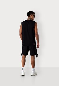 Schwarzes ärmelloses Oberteil und Shorts aus leichtem Stoff. Das Oberteil hat einen runden Halsausschnitt; die Shorts verfügen über Seitentaschen und verstellbare Bänder. Weiße Sneakers.