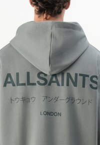 Hettegenser i lysegråt stoff. Har teksttrykk i mørkegrønn, inkludert "ALLSAINTS" og "LONDON" på baksiden.