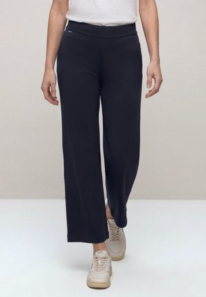 Person iført sorte bukser med vidde ben, hvide sneakers og en hvid skjorte, går fremad på en ensartet lys baggrund.