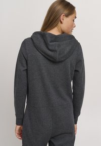 Grå hettegenser med myk tekstur. Har en tettsittende design, lange ermer og kengurulomme. Bakken viser subtile sømdetaljer.