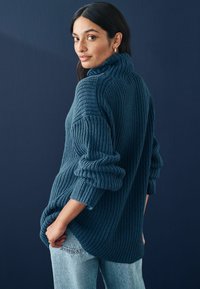 Pulover tricotat de culoare teal, cu guler înalt, textură ribbed și croială supradimensionată; asortat cu blugi deschiși, pe un fundal albastru închis.