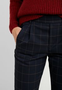 Personne portant un pantalon à carreaux bleu foncé avec une main dans la poche et un pull en tricot rouge.