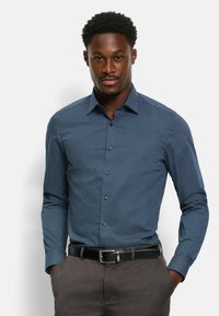 Hemd in dunkelblauem Stoff mit einem kleinen geometrischen Muster, langen Ärmeln und einem Kragen. Kombiniert mit grauen Hosen und einem schwarzen Gürtel.