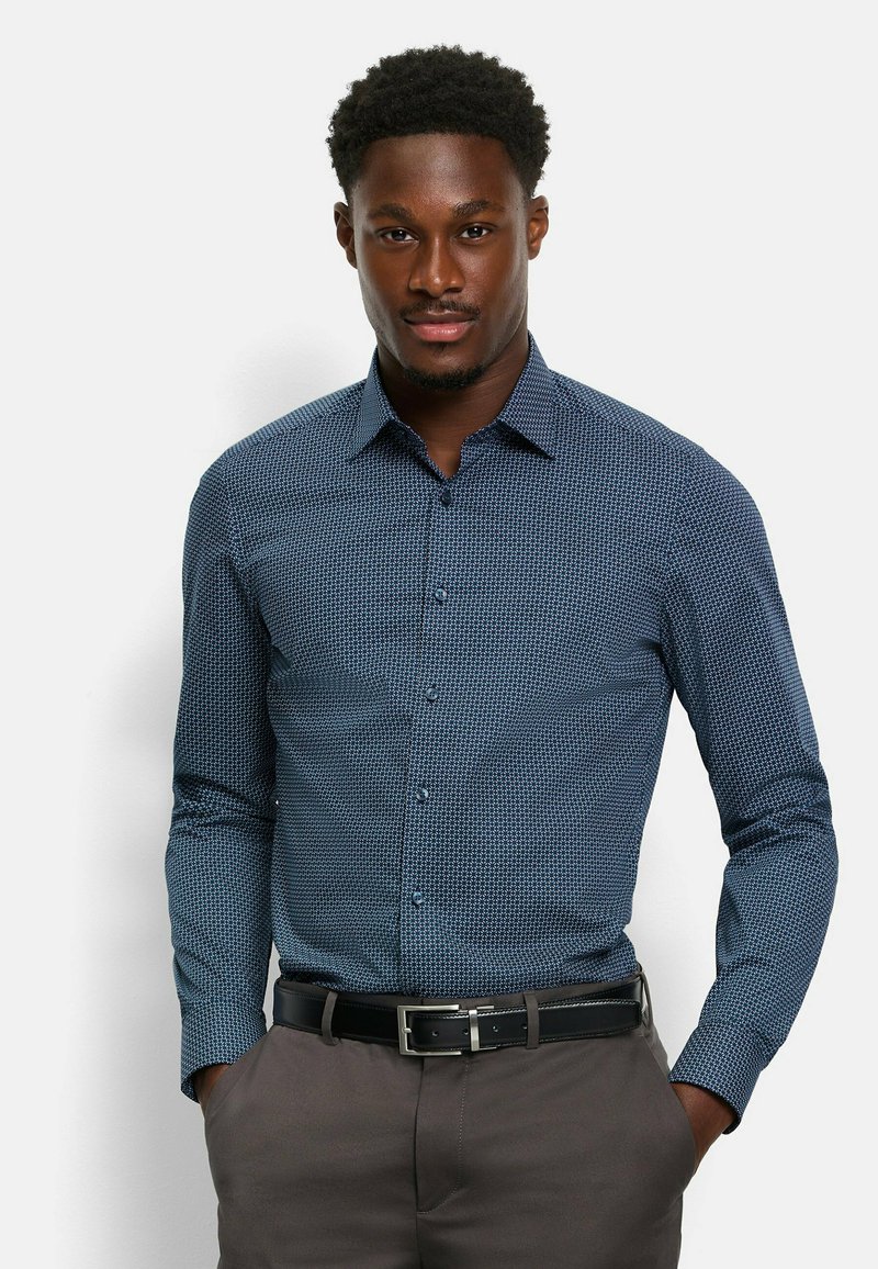 Hemd in dunkelblauem Stoff mit einem kleinen geometrischen Muster, langen Ärmeln und einem Kragen. Kombiniert mit grauen Hosen und einem schwarzen Gürtel.