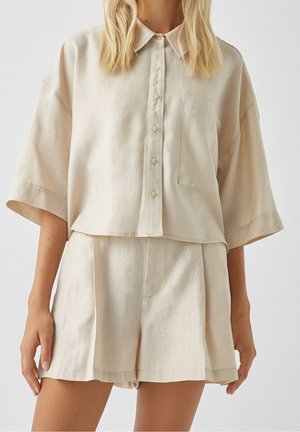 Femme portant une chemise beige ample à manches courtes boutonnée avec poche poitrine et un short assorti taille haute.