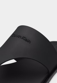 Sandale à glissière noire Calvin Klein avec large bride et nom de la marque embossé sur la partie supérieure, sur un fond uni.