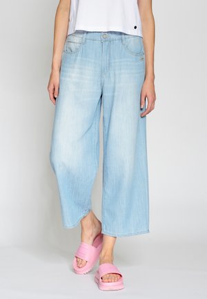Jeans de pierna ancha azul claro con bordes deshilachados y bolsillos frontales, combinados con sandalias chunky rosas. Textura de denim suave.