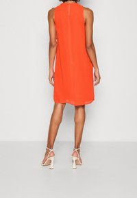 Robe orange sans manches avec une fermeture éclair au dos, présentant une coupe droite et une texture lisse. Portée avec des talons à lanières de couleur claire.
