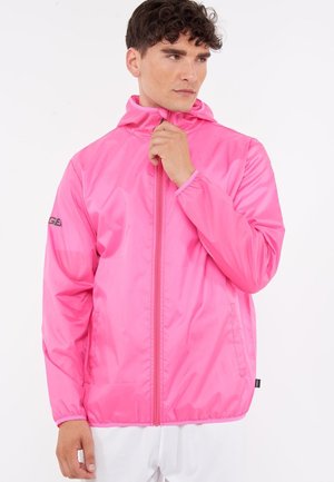 Man with curly hair wearing a bright pink hooded windbreaker jacket, partially zipped, looking to the side against a white background.