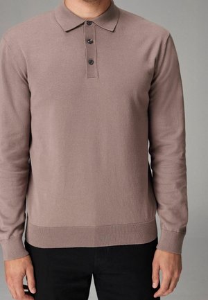 Mand iført en mauve langærmet polotrøje med tre knapper, kombineret med sorte bukser, stående mod en neutral baggrund.