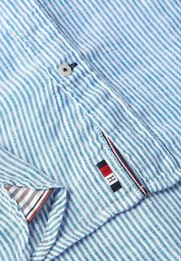 Blå och vit randig skjorta, tillverkad av lättviktig tyg. Har en knäppning med knappar och ett märke med röda och svarta accenter.