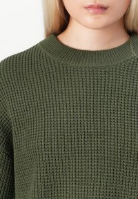 Κοντινό πλάνο ατόμου που φοράει πλεκτό πουλόβερ ελιάς με ριμπ λεπτομέρεια στο στρογγυλό λαιμό, δείχνοντας το κάτω μέρος του προσώπου και το λαιμό.