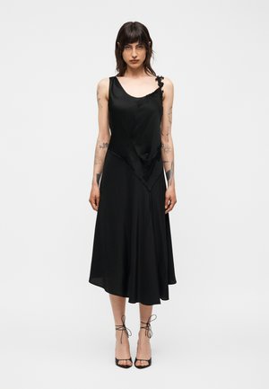 Kobieta z tatuażami, ubrana w czarną asymetryczną sukienkę midi bez rękawów i czarne szpilki z paskami, stojąca na jednolitym białym tle.