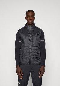 Melna izolēta veste ar augstu apkakli, ar zipa aizdari priekšā, sānu kabatām un logo detaļu. Teksturēta auduma ar gludu apdari.