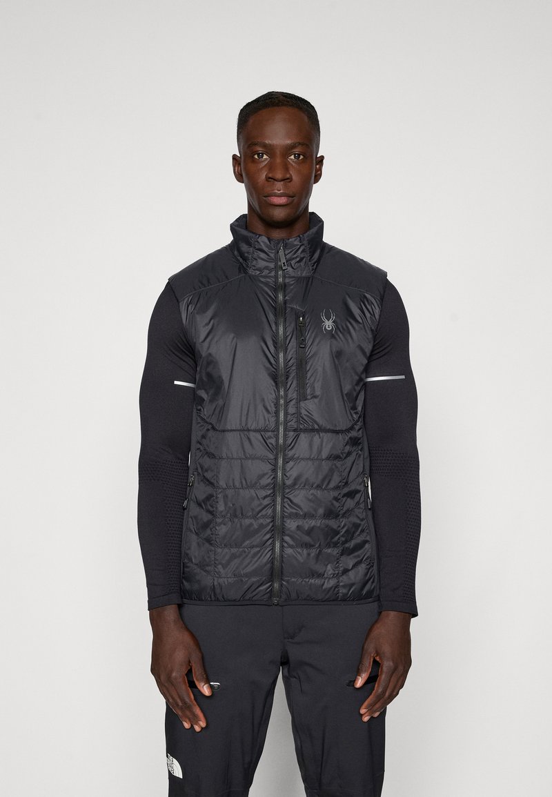 Melna izolēta veste ar augstu apkakli, ar zipa aizdari priekšā, sānu kabatām un logo detaļu. Teksturēta auduma ar gludu apdari.