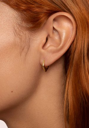 Goldene geometrische Creolen, die am Ohr eines Modells präsentiert werden, mit einem schlanken, kantigen Design und einer polierten metallischen Oberfläche.