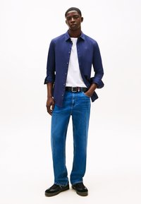 Camisa azul-marinho de botões sobre t-shirt branca, jeans azul de corte solto, cinto preto e ténis pretos. Design simples, textura suave, aspeto casual.