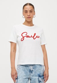 White cotton t-shirt featuring red fluffy "Smile" text. Short sleeves and a relaxed fit. Paired with light blue denim pants.