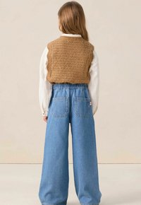 Gilet en tricot marron avec un bord festonné, porté par-dessus une chemise blanche à manches longues ; associé à un pantalon en denim bleu à jambes larges avec des poches arrière.