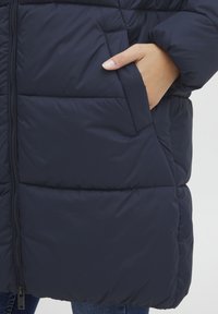 Veste en doudoune marine avec un design matelassé, fermeture éclair et poches latérales. Le matériau semble lisse et rembourré pour une isolation optimale.