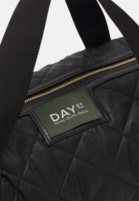 Sac noir matelassé en nylon avec une étiquette verte portant l'inscription "DAY ET" et une fermeture éclair dorée. Tissu solide et texturé avec des bandoulières contrastantes.