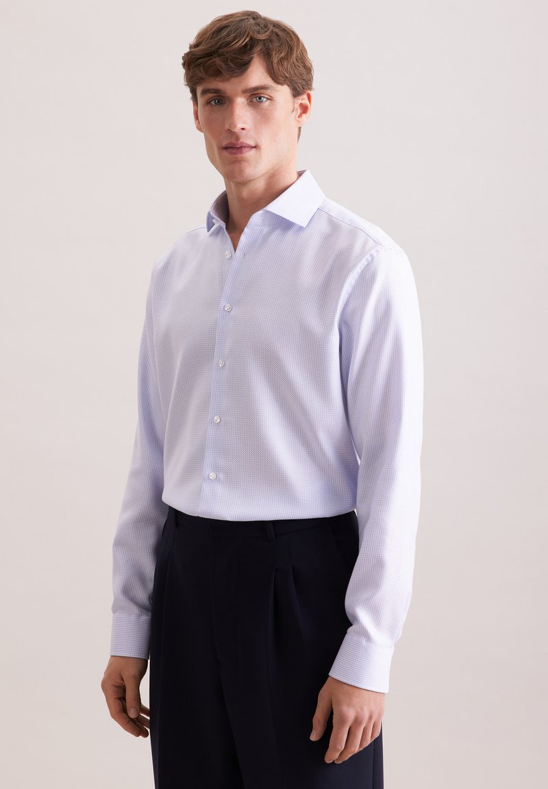 Jeune homme portant une chemise habillée texturée bleu clair rentrée dans un pantalon noir plissé, debout devant un fond uni.