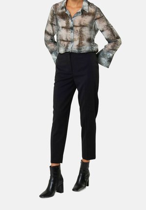 Kobieta ubrana w przezroczystą, zapinaną na guziki bluzkę z długim rękawem z abstrakcyjnymi wzorami, czarne skrócone spodnie i czarne botki na obcasie, stojąca.