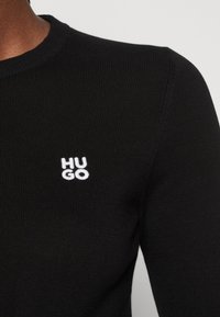 Černý žebrovaný pletený svetr s kulatým výstřihem a bílým vyšitým logem "HUGO" na levé části hrudníku.