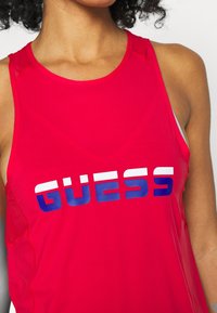Czerwona sportowa koszulka bez rękawów z wyraźnym logotypem "GUESS" w białym i niebieskim kolorze. Materiał jest lekki o gładkiej fakturze.