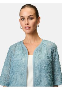 Hellblauer floraler Spitzen-Cardigan mit kurzen Ärmeln, strukturiertem Design und zwei vorderen Taschen, verziert mit einer dekorativen Borte entlang der Kanten.