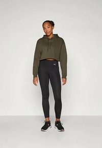 Žena stojí pred jednoduchým pozadím a má na sebe olivovozelenú skrátenú mikinu, čierne legíny Puma a čierne športové topánky s ružovými akcentmi.