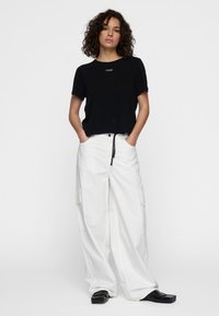 Zwart cropped t-shirt met klein logo, gecombineerd met witte cargobroek met wijde pijpen, zijzakken en een trekkoord in de taille. Zwarte schoenen maken de outfit af.