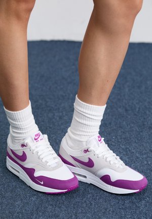 Un paio di sneakers Nike Air Max bianche e viola indossate con calze bianche a crew su un pavimento inmoquette blu.