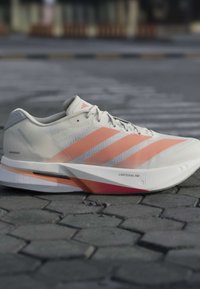 Λευκό και πορτοκαλί αθλητικό παπούτσι Adidas σε γκρι πλακόστρωτο, με τρεις πορτοκαλί ρίγες και "LIGHTSTRIKE PRO" στη σόλα.