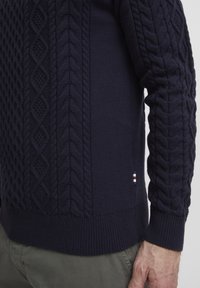 Pull en maille navy avec des motifs en câble et en losange, poignets et ourlet côtelés, avec un petit logo sur le côté.