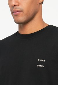 T-shirt preta em algodão com decote redondo e dois pequenos logótipos bordados em branco "SAMSØE" na área do peito esquerdo.
