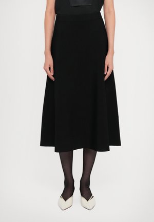 Woman wearing a black midi skirt, black tights, white pointed-toe heels, standing against a plain white background.