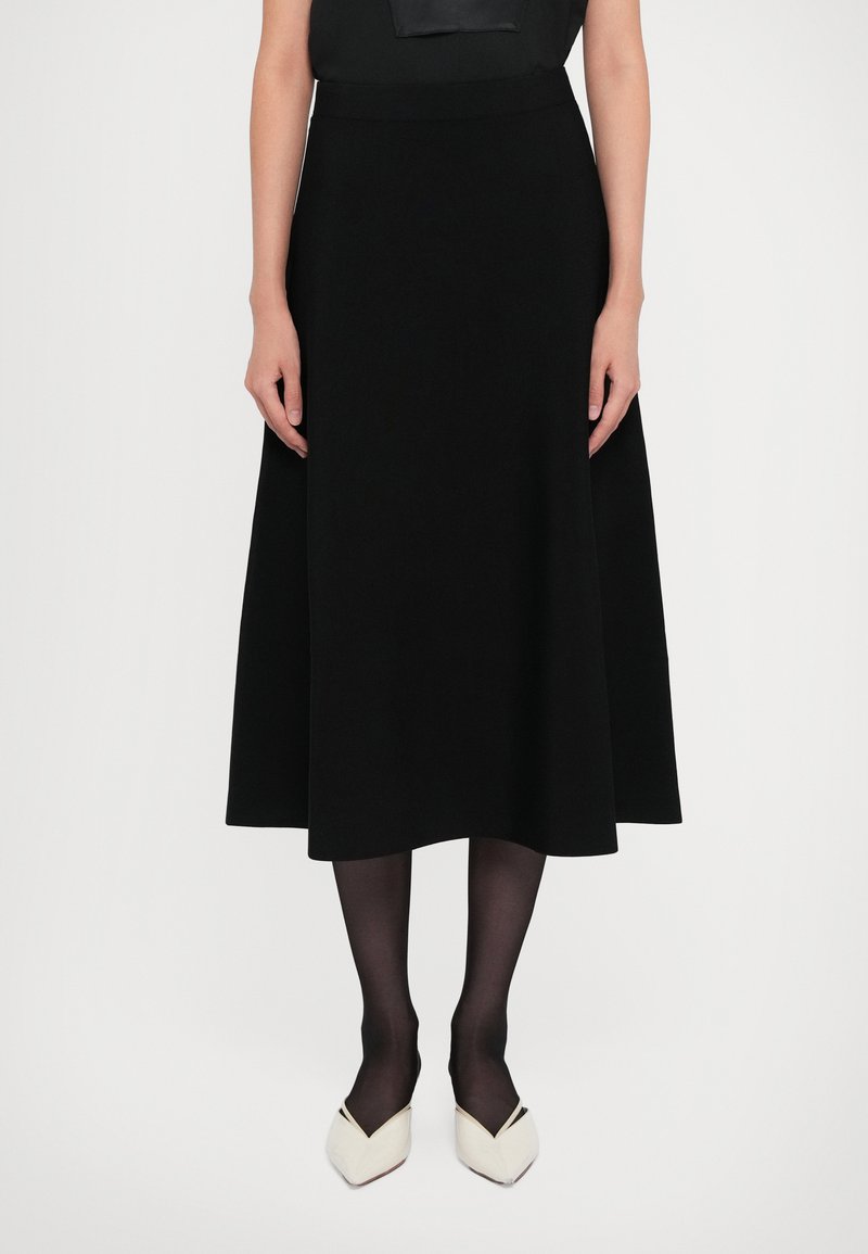 Kvinna som bär en svart midikjol, svarta leggings, vita spetsiga klackskor, stående mot en enkel vit bakgrund.