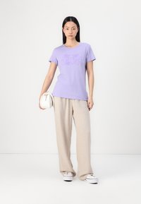 Lila crew neck t-paita, jossa on painettu logo, yhdistettynä vaaleanbeigeihin leveälahkeisiin housuihin ja valkoisiin tennareihin. Malli pitää valkoista laukkua.