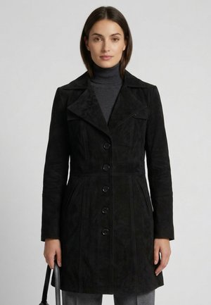 Vrouw draagt een zwarte jas met knopen en een grijze coltrui, houdt een zwarte handtas vast, tegen een effen lichtgrijze achtergrond.