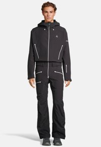 Giacca nera impermeabile con cappuccio regolabile e tasche con zip. Abbinata a pantaloni neri coordinati con dettagli con zip e una vestibilità slim.