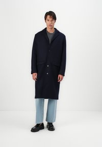 Cappotto in misto lana navy con colletto a punta, chiusura a cinque bottoni e tasche frontali, abbinato a jeans azzurri a gamba dritta e scarpe nere.