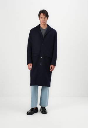 Marineblauer Wollmischmantel mit Reverskragen, Fünf-Knopf-Verschluss und Fronttaschen, kombiniert mit hellblauen, geraden Jeans und schwarzen Schuhen.