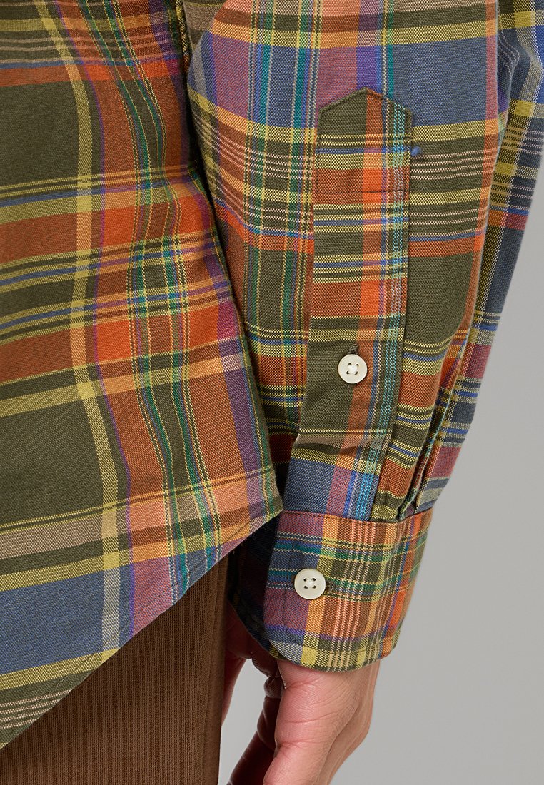 Chemise en coton à carreaux ; présente des motifs orange, vert et bleu. Comprend un poignet à bouton unique et un col rabattu sur un fond beige.